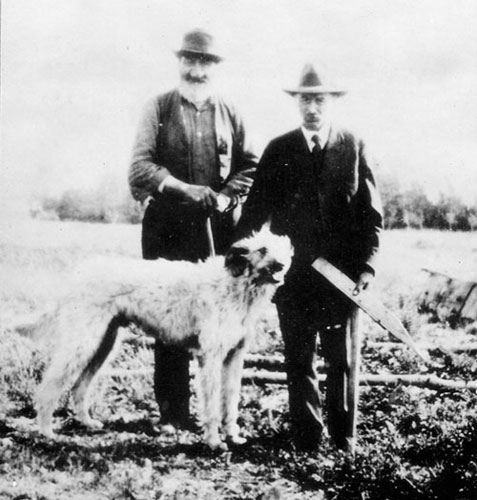 A black and white photograph of two men standing in a field with a dog. Peter Erasmus on the left has a bushy white beard and a distinctively misshapen nose.